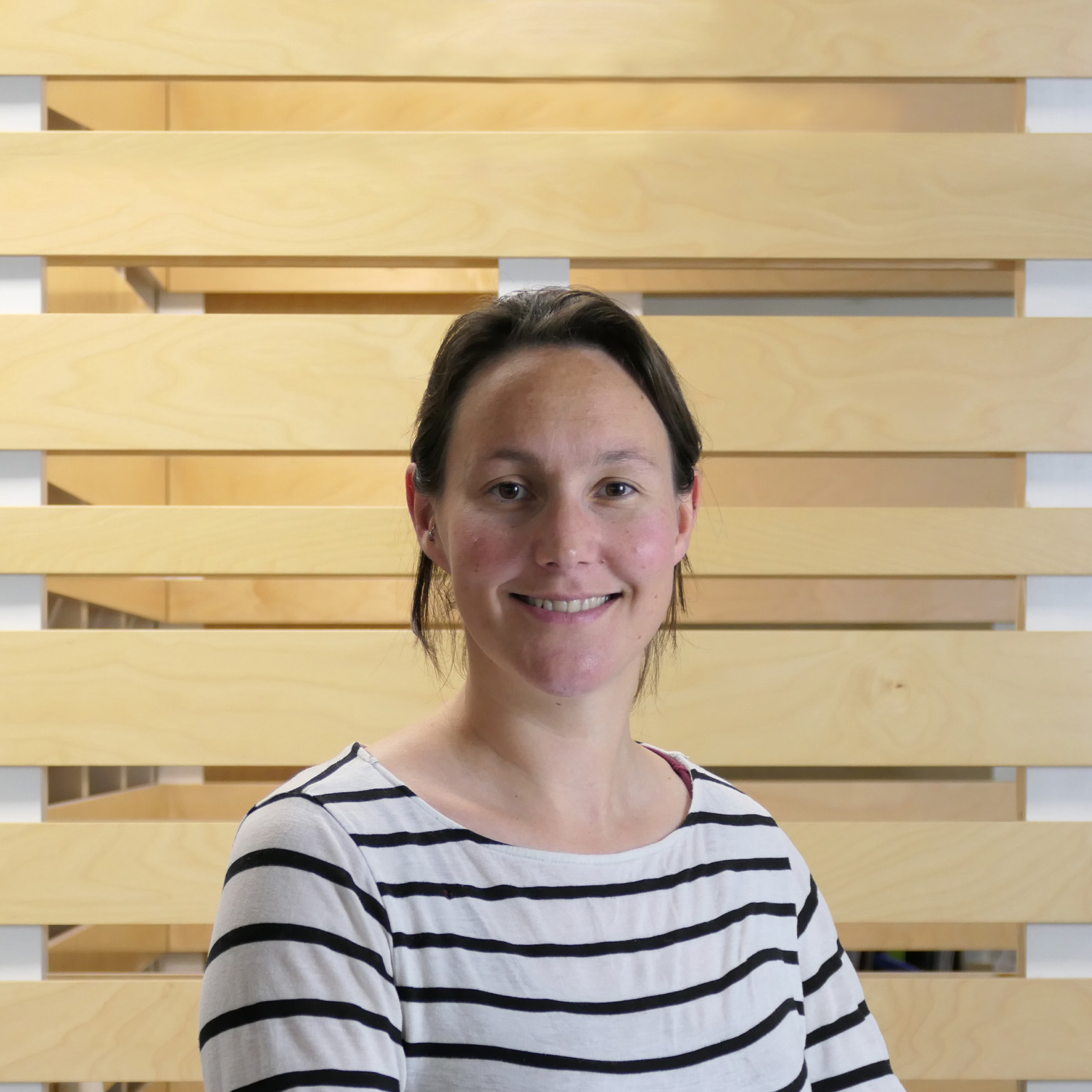 Elspeth Kirkman wearing a white shirt with black stripes, smiling against a backdrop of wood paneling.