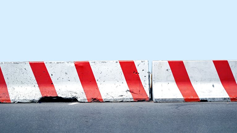 A pair of red and white concrete road barriers aligned on a street against a light blue background. 