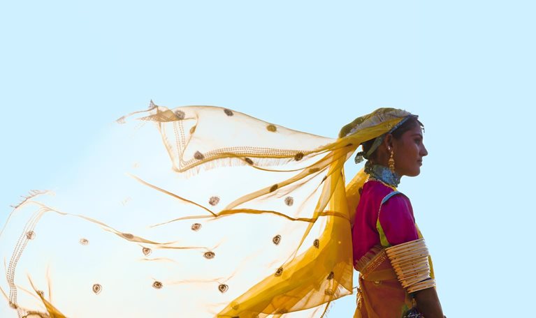 A woman from India stands tall and proud in a vibrant saree, her yellow veil fluttering in the wind against a backdrop of a clear blue sky.