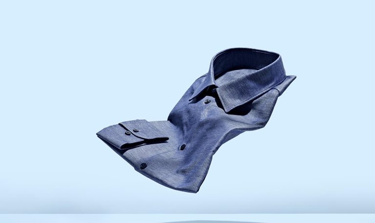 A chambray mens dress shirt floating weightlessly against a light blue background.