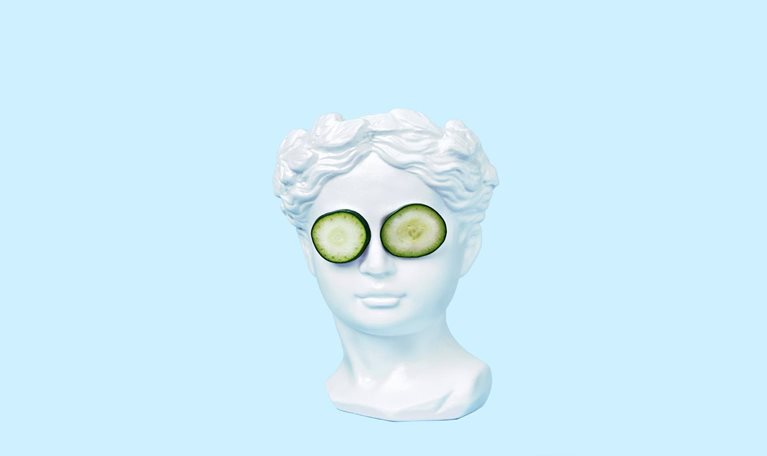 A bust of a young woman, inspired by ancient Greek art, wearing cucumber slices on her eyes and set against a light blue backdrop.