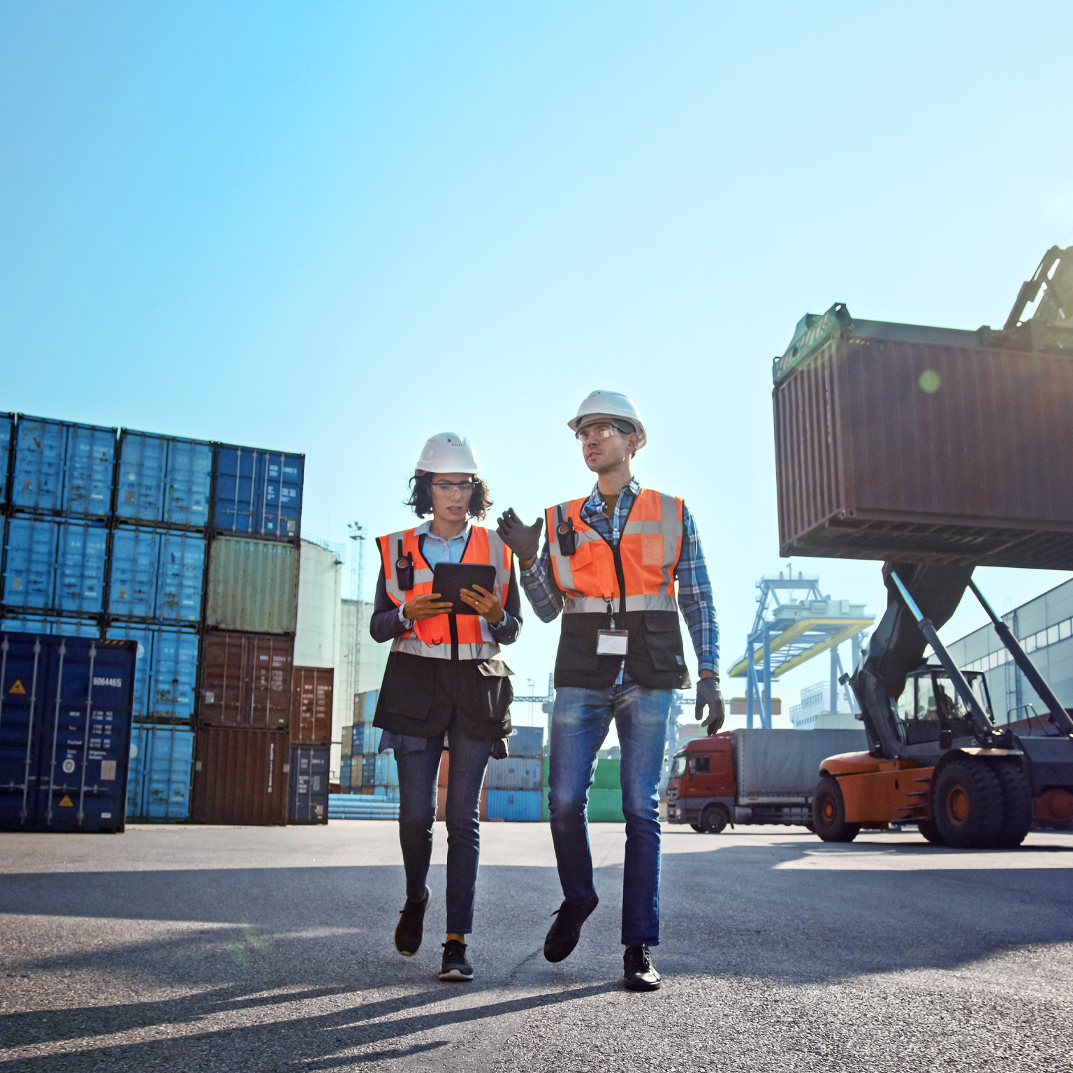 Female industrial engineer with tablet computer and male foreman worker in hard hats and safety vests walk in container terminal