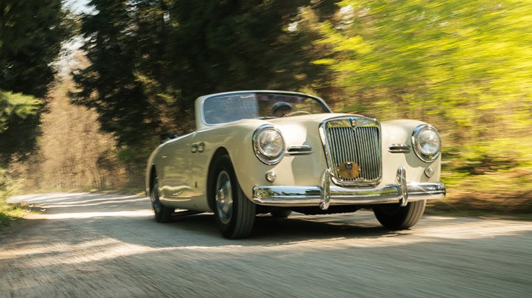 Classis car driving along country road through woods.