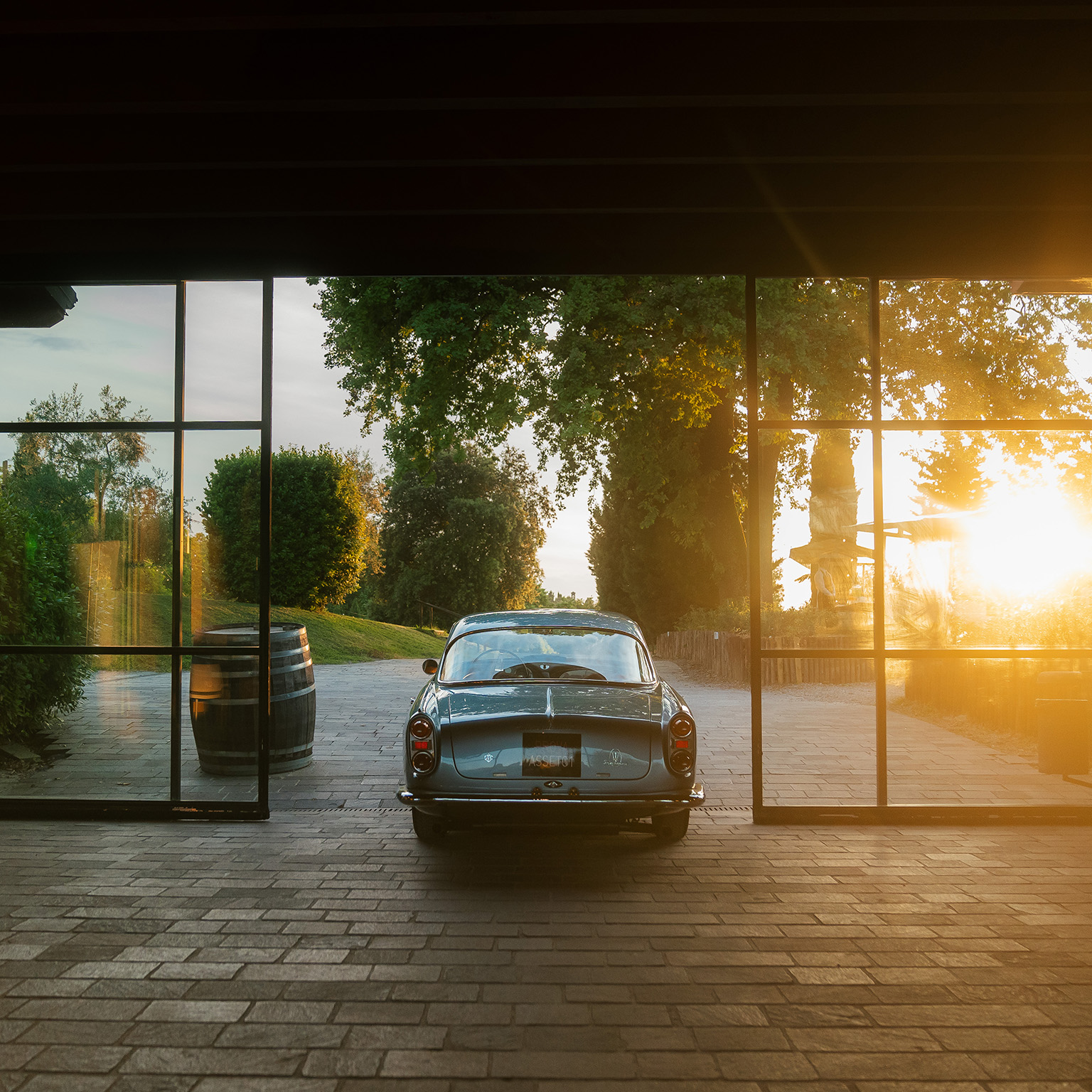 Rear view of classic car driving out from behind glass garage doors as sun sets in background.