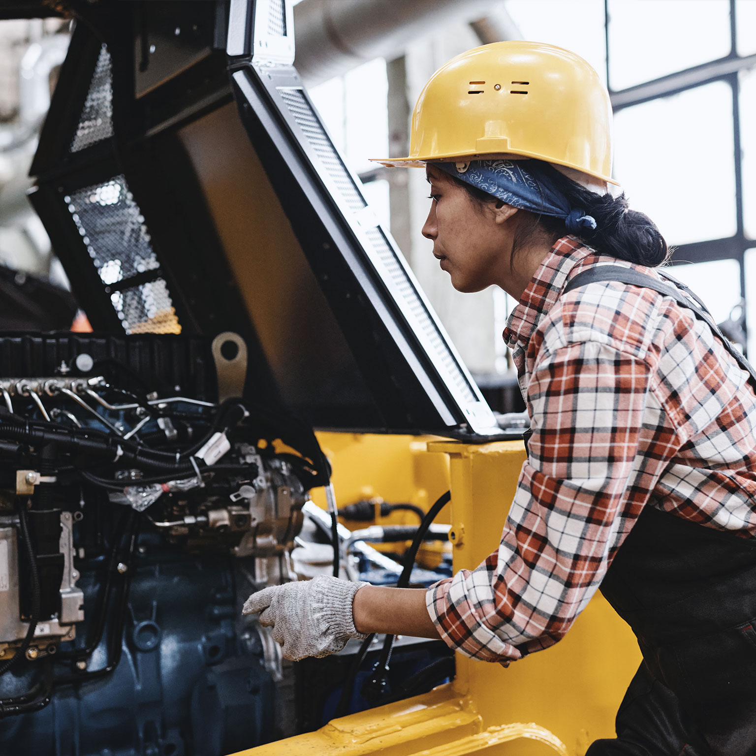 Young female technician in coveralls and hardhat checking motor of machine