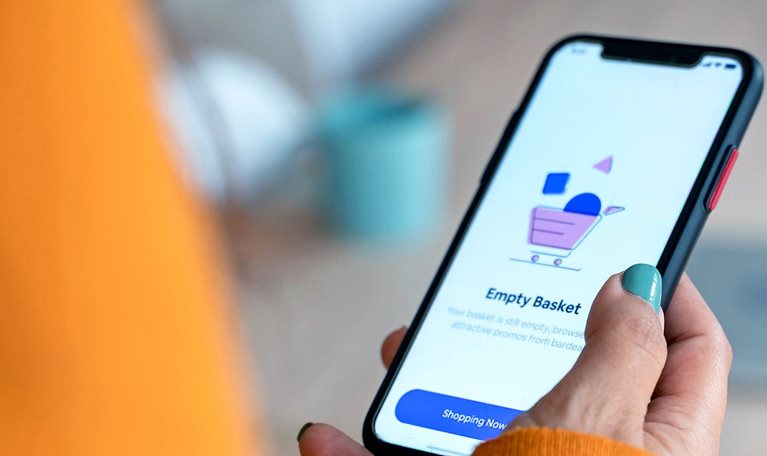 Person in an orange sweater holding a smartphone displaying a shopping app screen that says “Empty Basket”