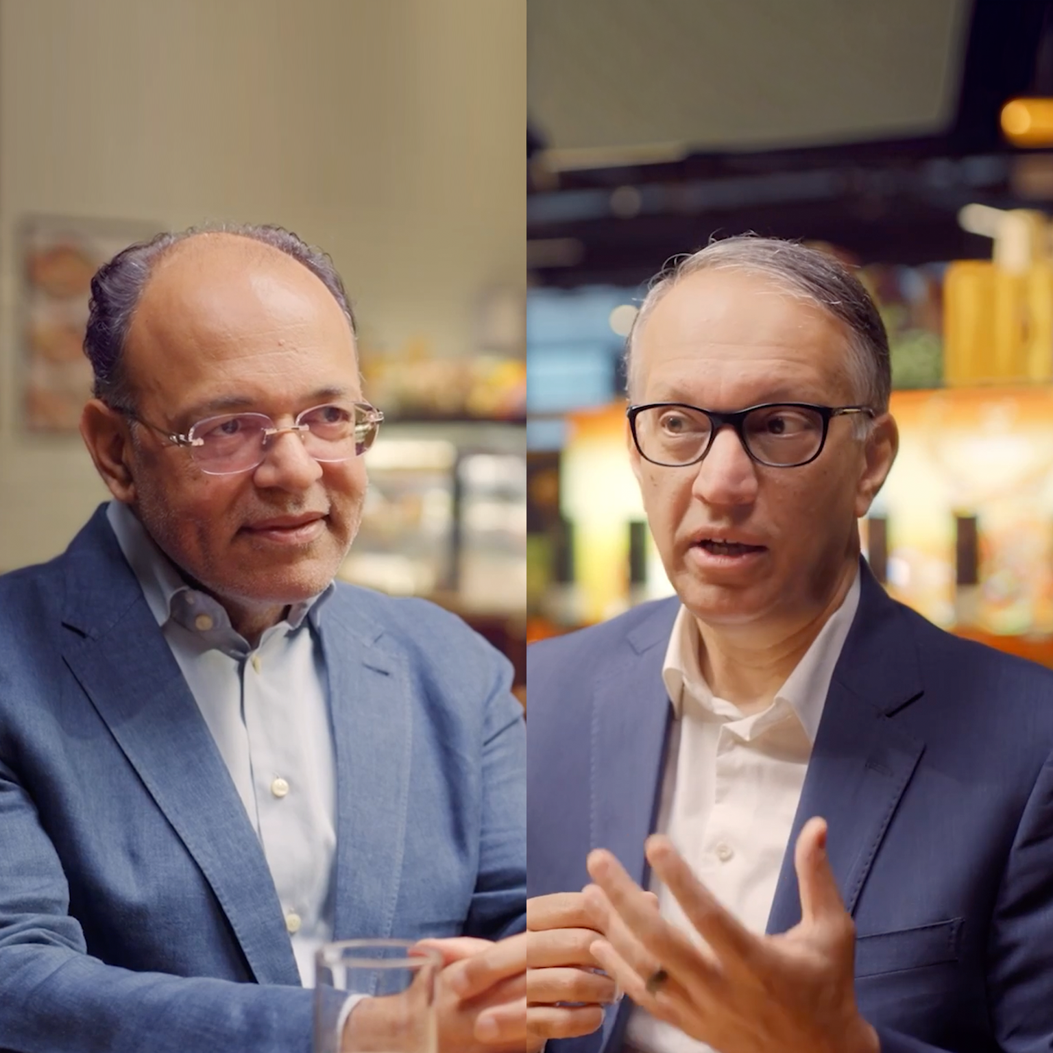 Vipul Chawla in a blue suit and glasses sits in a warmly lit store or café setting, smiling slightly, with shelves and lights blurred behind.