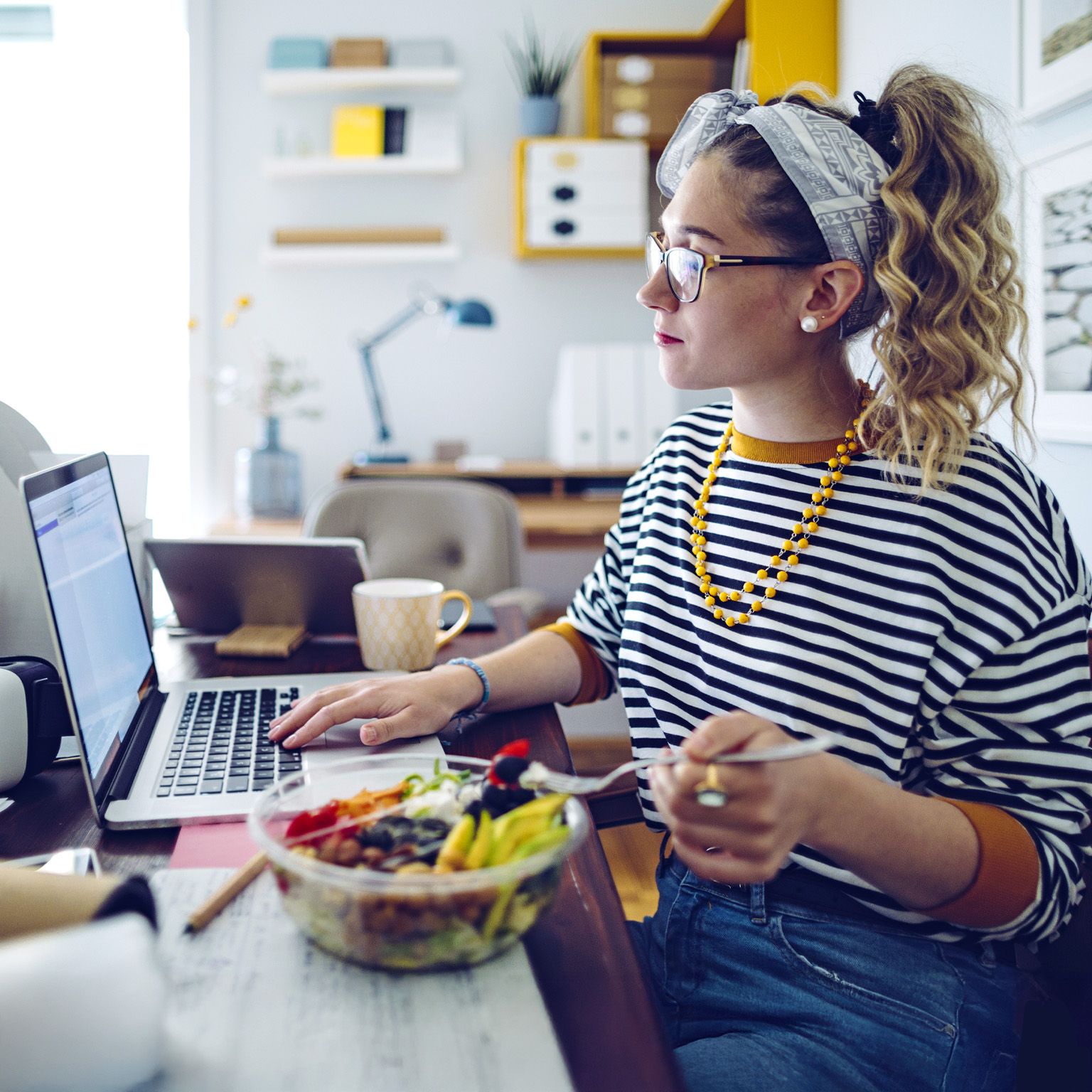 A woman wearing glasses and a striped shirt is seated at a desk, looking at her laptop and eating a salad.