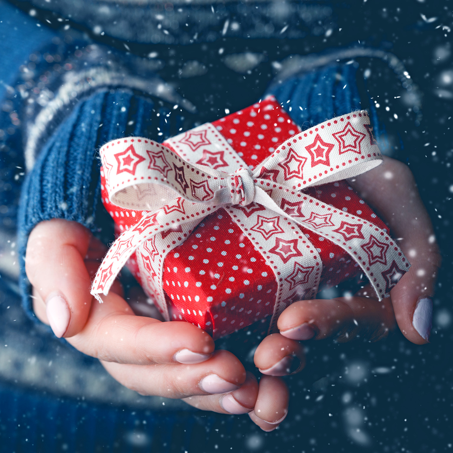 Close-up image of hands holding a small red gift box with a festive white bow with stars on it, with snow falling in the background.