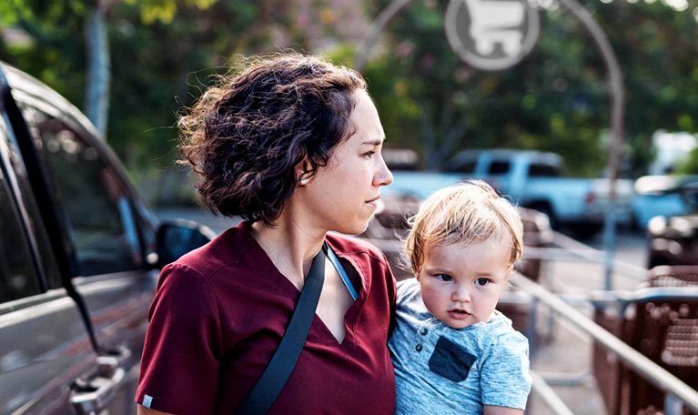 A woman holding a young child in a parking lot near shopping carts and parked cars. The woman looks off into the distance with a focused expression, while the child looks toward the camera.
