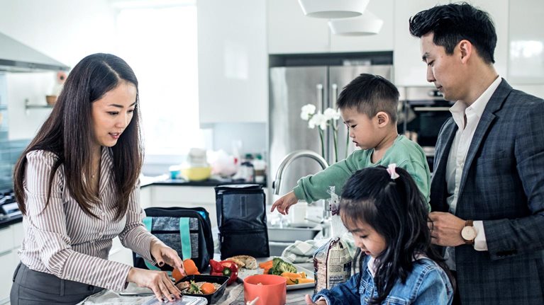 A family gathered in a bright modern kitchen, with parents and two children preparing food and packing containers together.