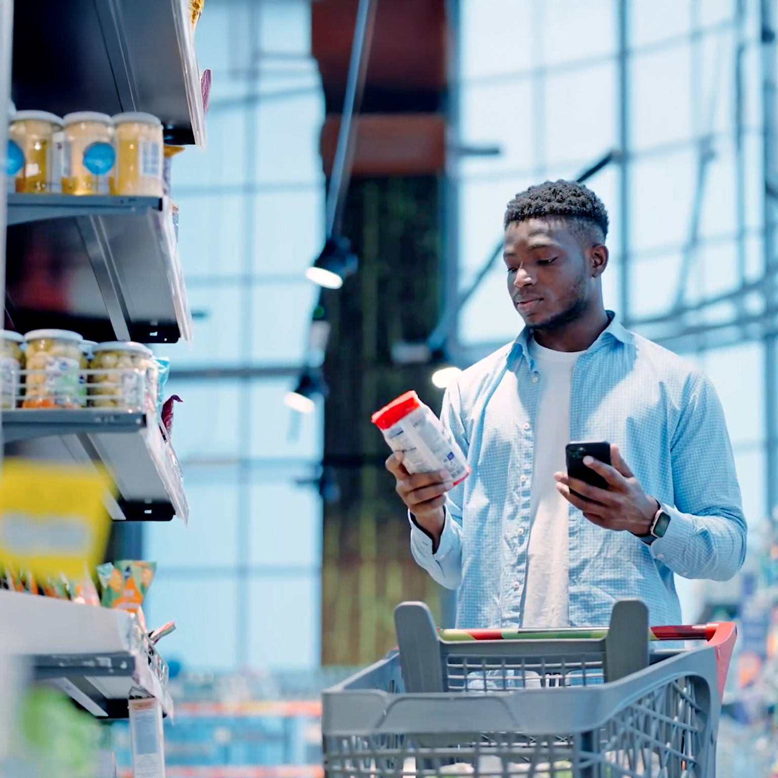 Person in a grocery store comparing a product label while holding a smartphone next to a shopping cart.