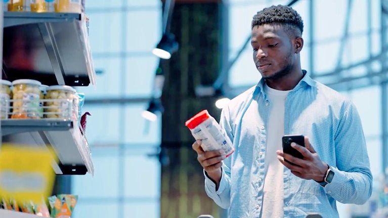 Person in a grocery store comparing a product label while holding a smartphone next to a shopping cart.