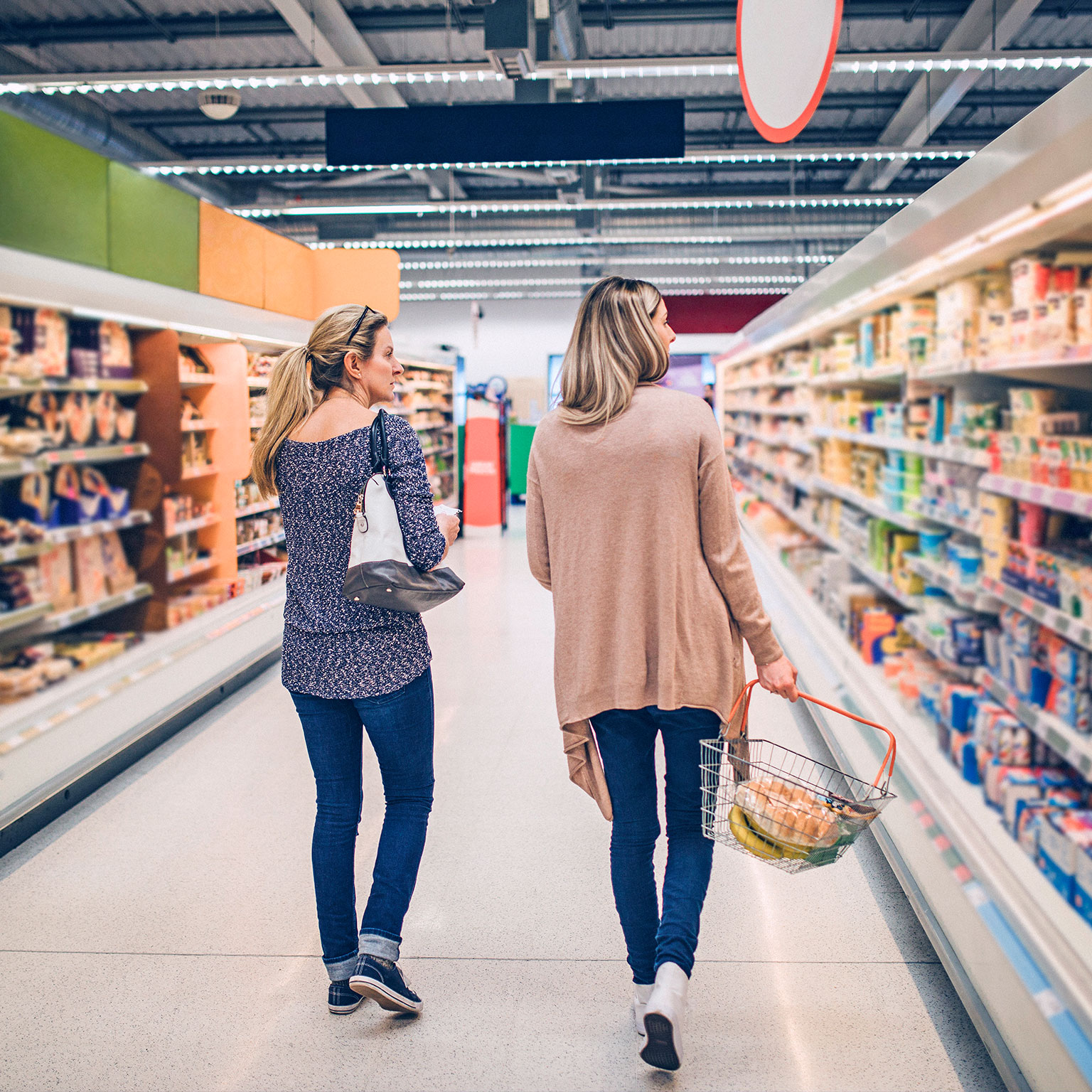 Two women walking down a grocery store aisle with a shopping basket