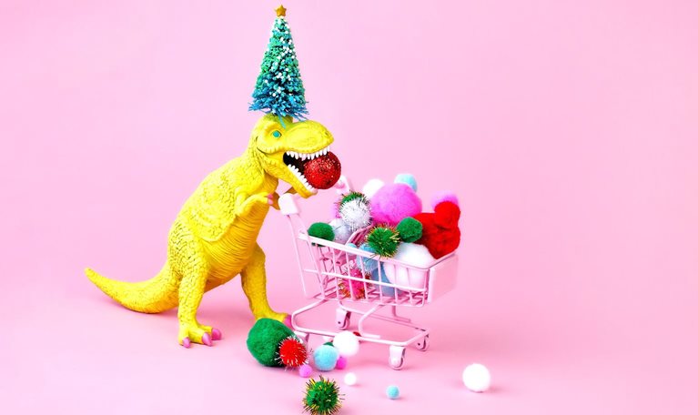 A festive image of a toy dinosaur wearing a Christmas tree on its head and filling a shopping cart with multicolored puff balls.