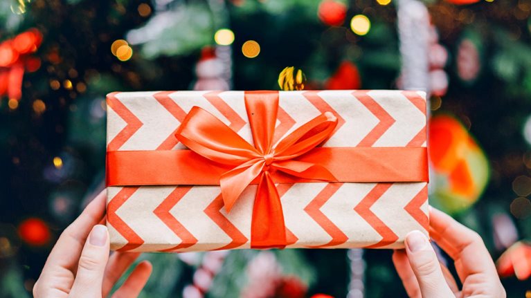 A pair of hands holding a wrapped gift box decorated with a red chevron pattern and tied with a bright red ribbon. In the background, colorful lights and ornaments create a festive, holiday atmosphere.
