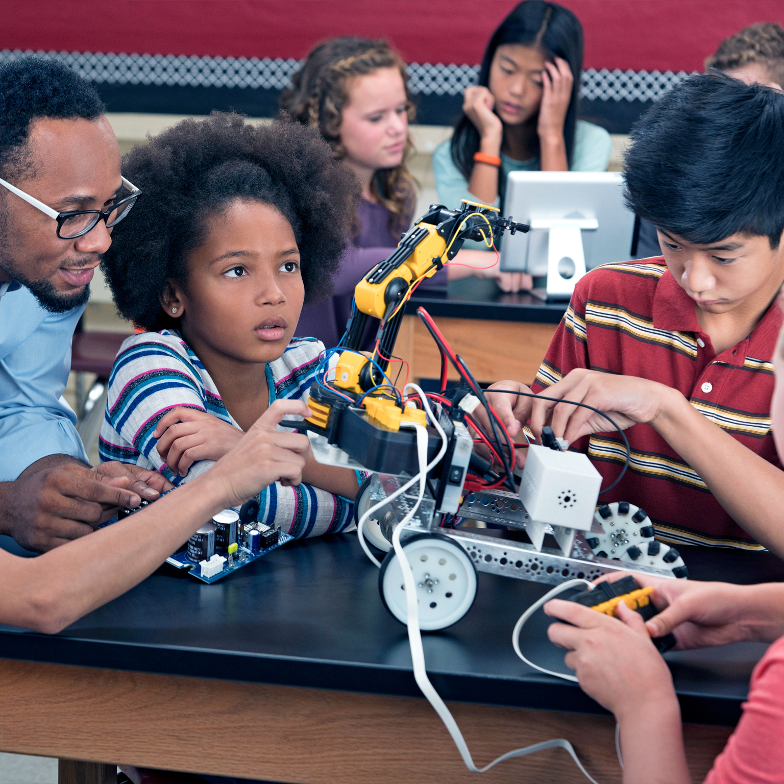 Children from diverse racial identities, who appear to be in middle school, are creating a mechanized forklift type of machine on wheels in a mechanics class with a teacher helping them. 