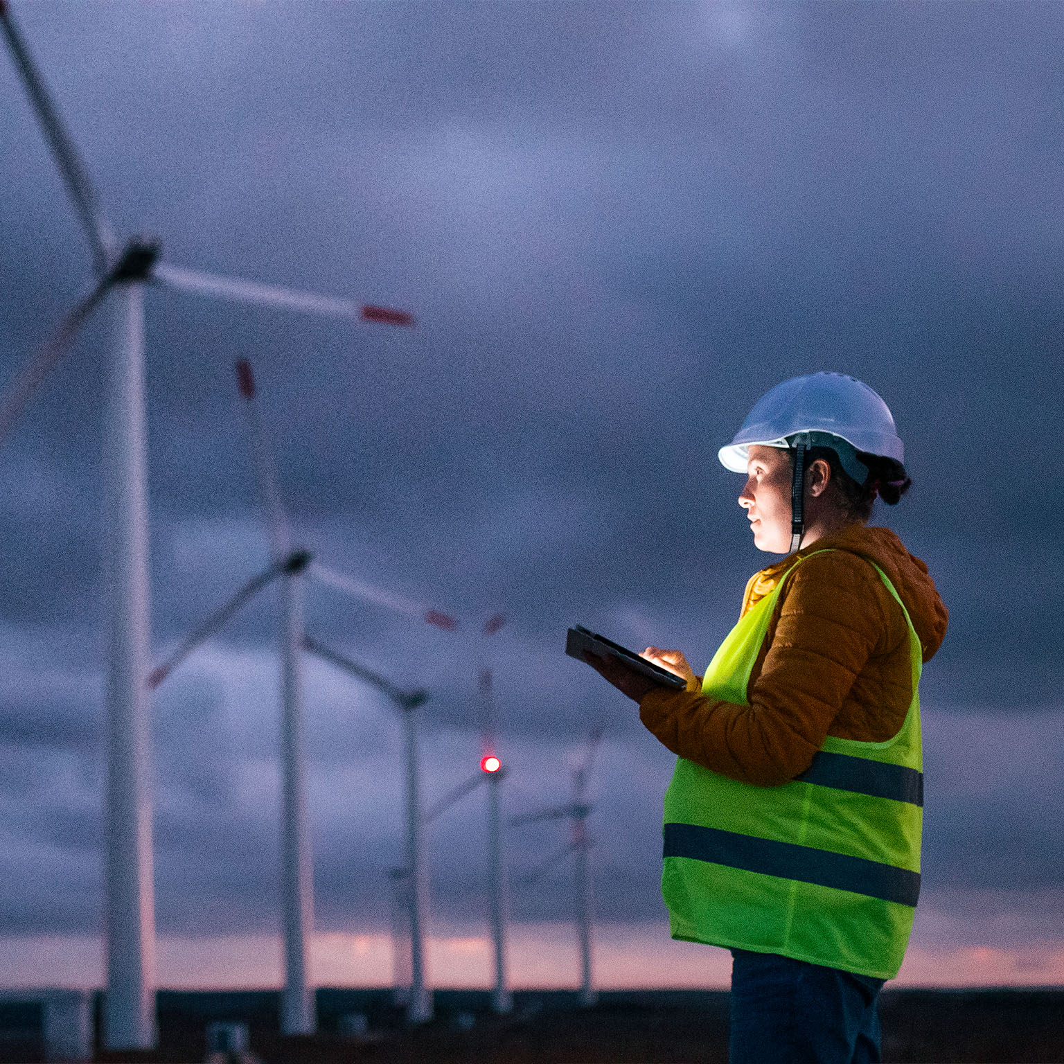 Renewable Energy Systems. Electricity Maintenance Engineer working on the field at a Wind Turbine Power station at dusk with a moody sky behind. Blurred motion. - stock photo