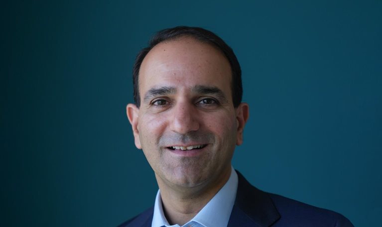 Anant Bhalla wearing a dark suit and a blue dress shirt. His face bears a light smile as he poses against a blue backdrop.