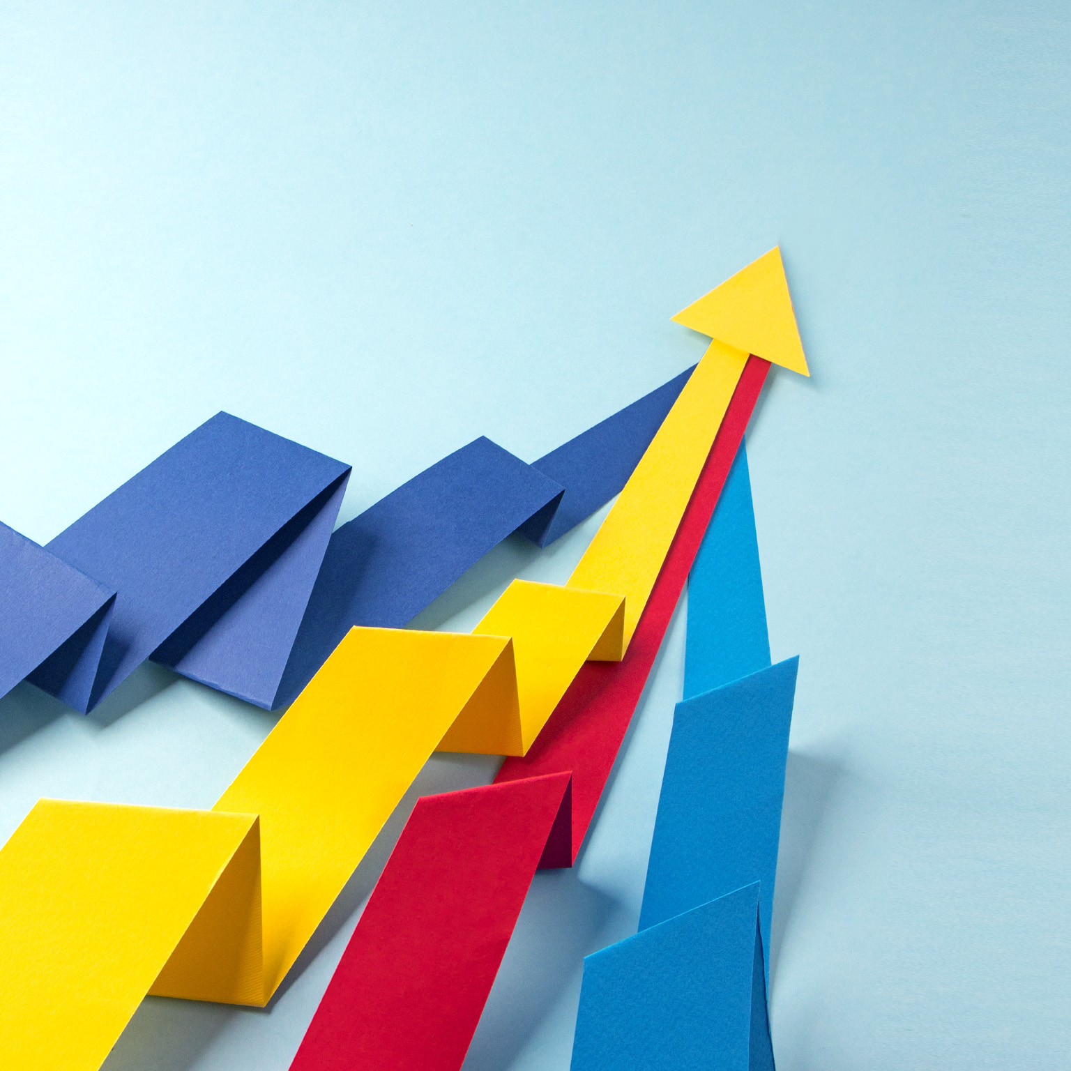 Red, blue, yellow paper arrows folded to appear like three-dimensional graphs merging at the top, with the yellow arrow head visible over others.