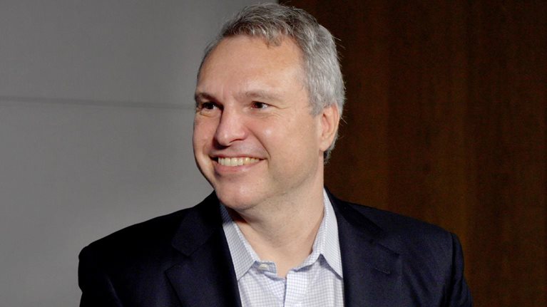 A portrait of Derek Waldron wearing a blue suit jacket over a collared shirt. He is smiling and looking to his right.