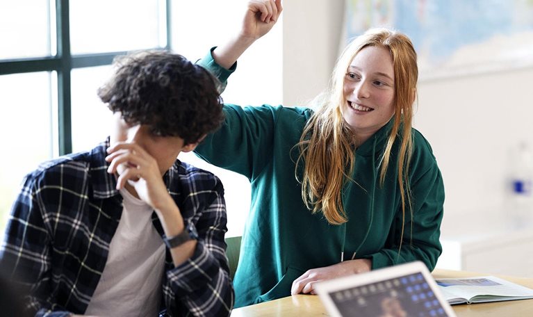 A group of students sitting at a classroom table, with a red-haired student in a green hoodie smiling and raising her hand while others, including one with a laptop, also participate.
