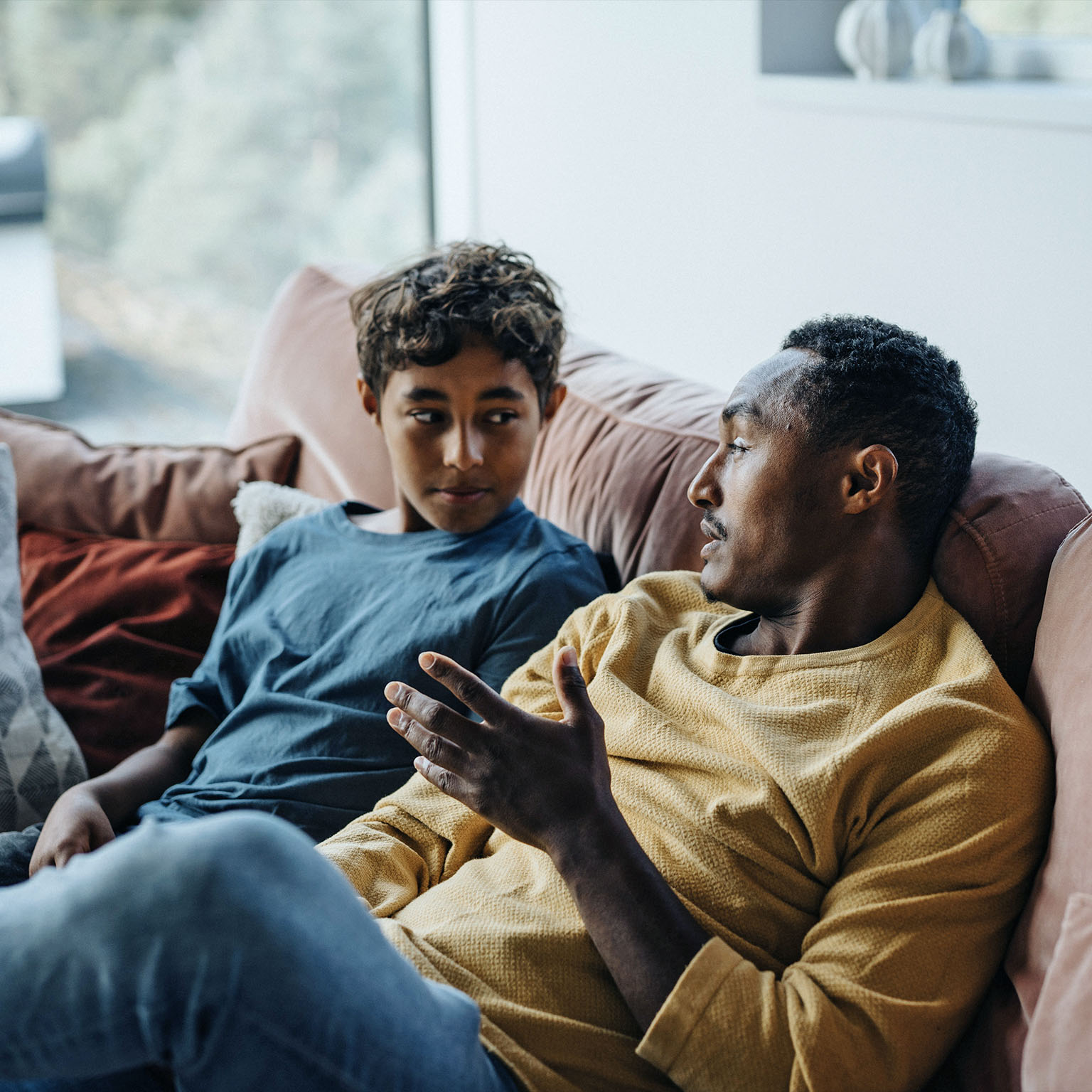 A man and a young boy sit together on a couch at home, facing each other as they talk, with the man gesturing while the boy listens attentively.