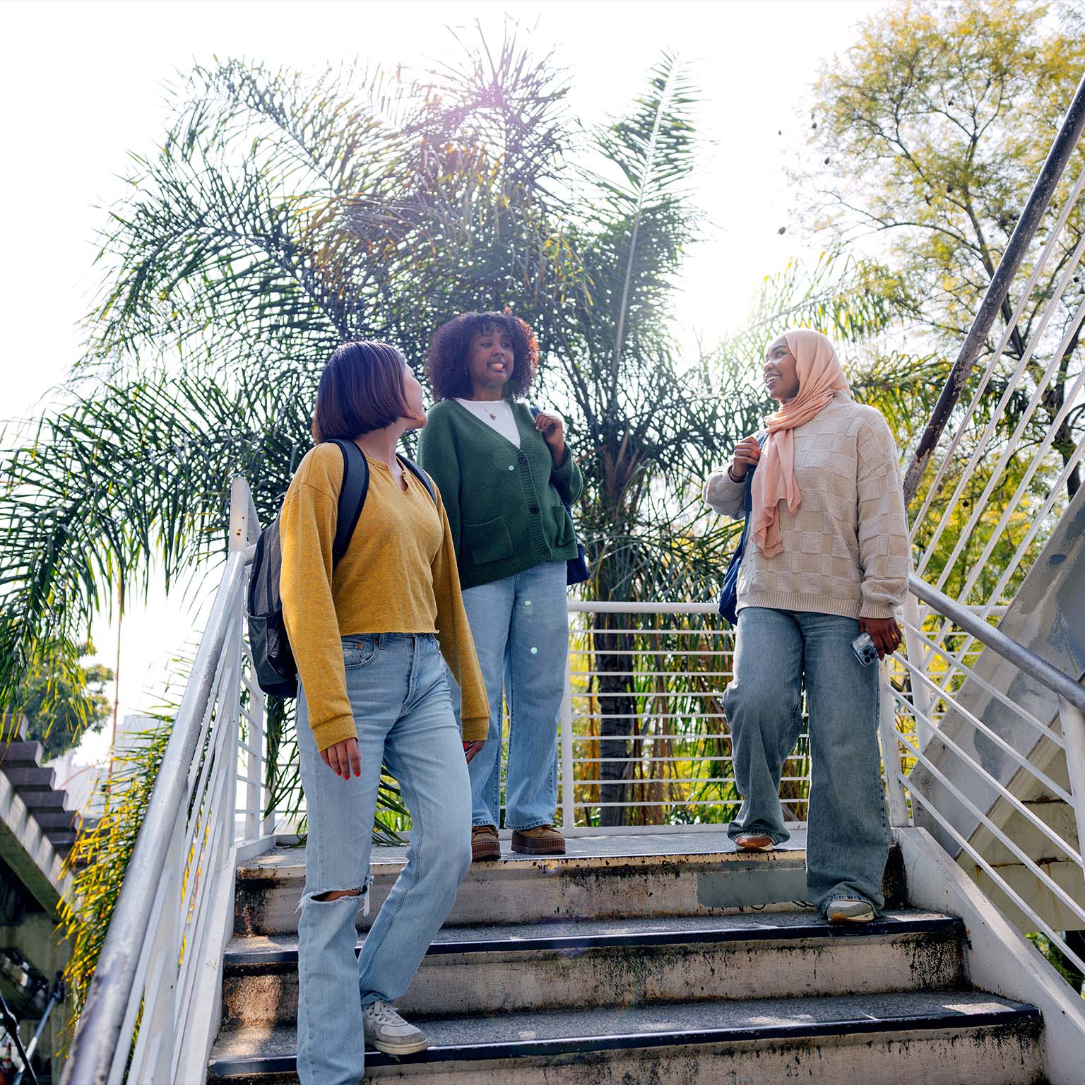 Three young women with backpacks walk down outdoor steps together, chatting and smiling, with palm trees and bright sunlight in the background.
