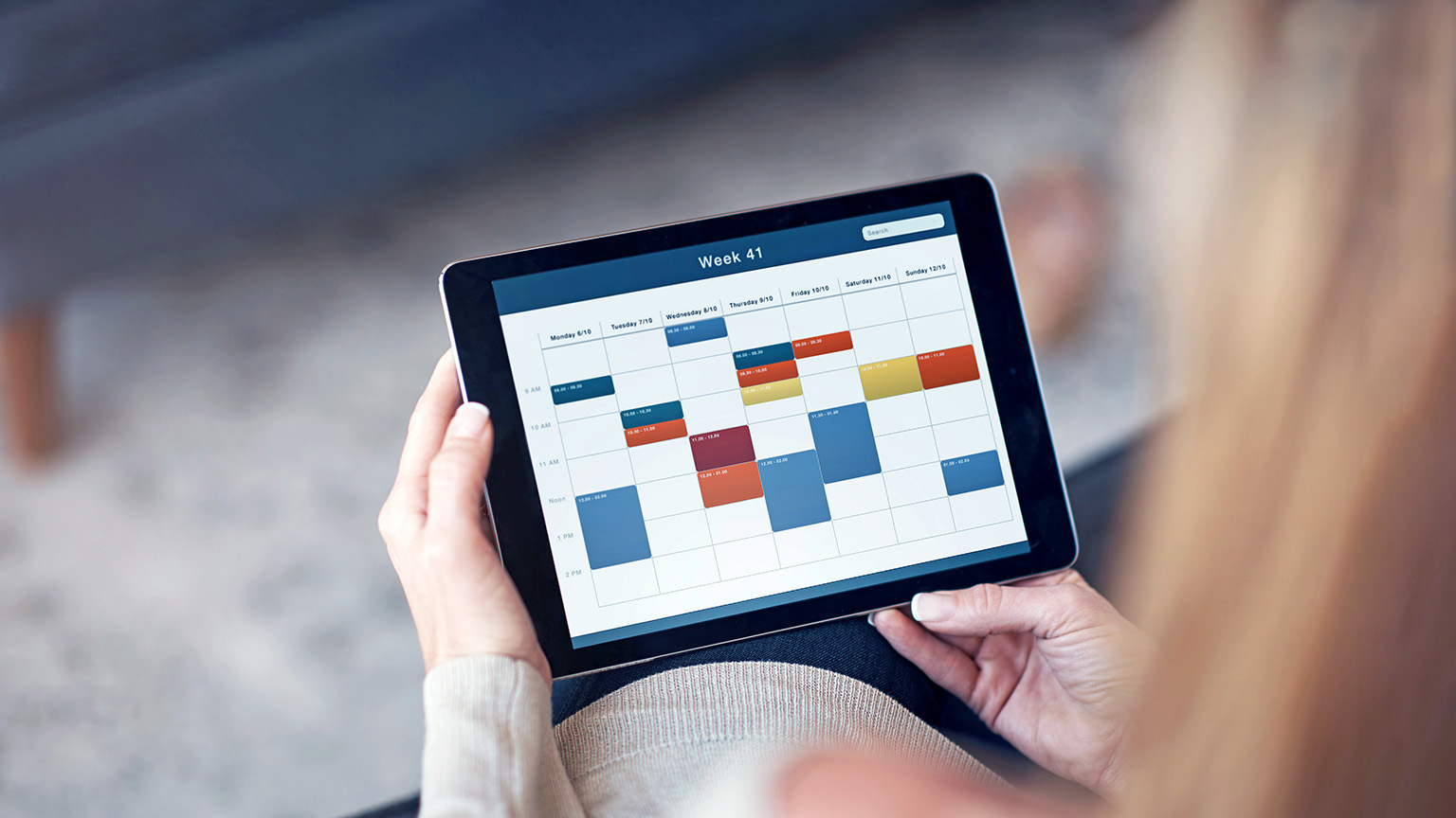 Cropped detail of A close-up view of a woman's hands using a tablet to access a scheduling calendar in a living room.