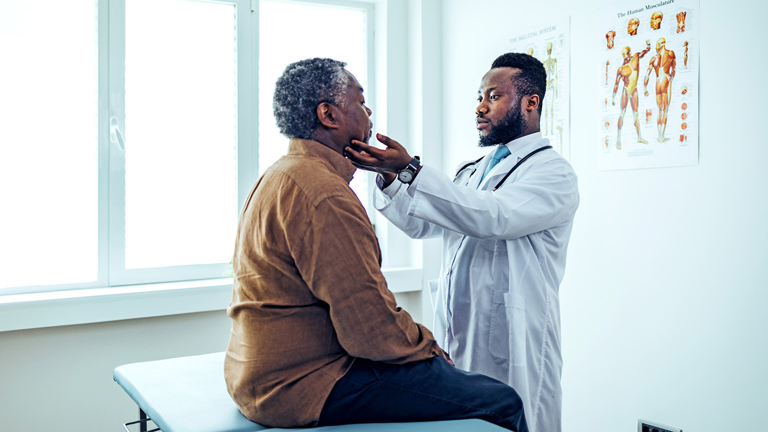 A mature patient is sitting on an exam table in a well-lit medical office while a doctor thoughtfully examines his lymph nodes by palpation.