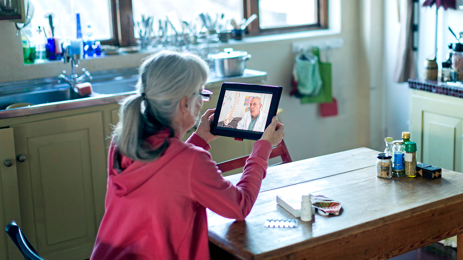 The doctor converses with a mature woman, her gray hair pulled back in a ponytail, as she sits at a kitchen table and talks through a tablet.