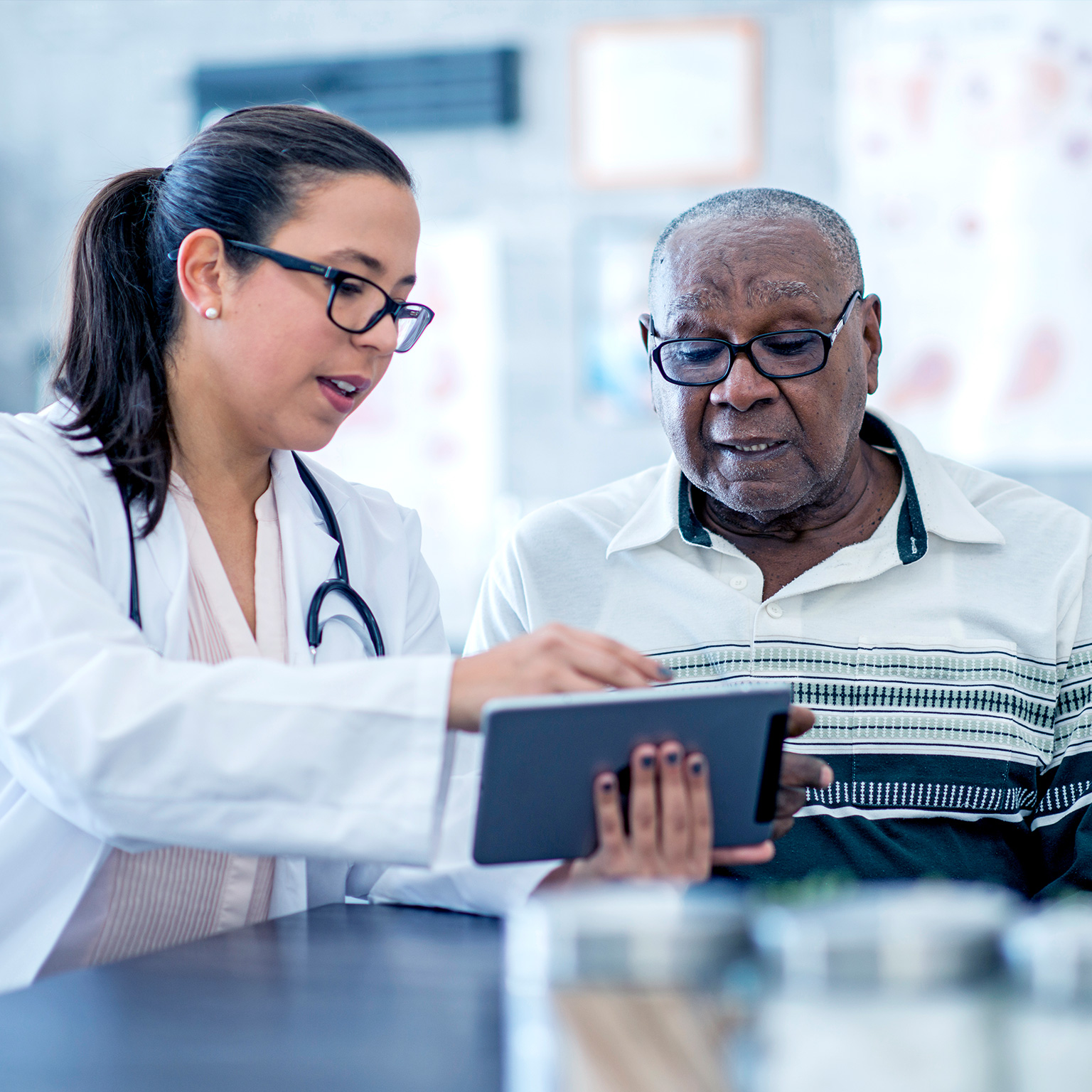 A doctor in white coat wearing a stethoscope, who is a woman, appears to explain something from an electronic tablet to an elderly black man.