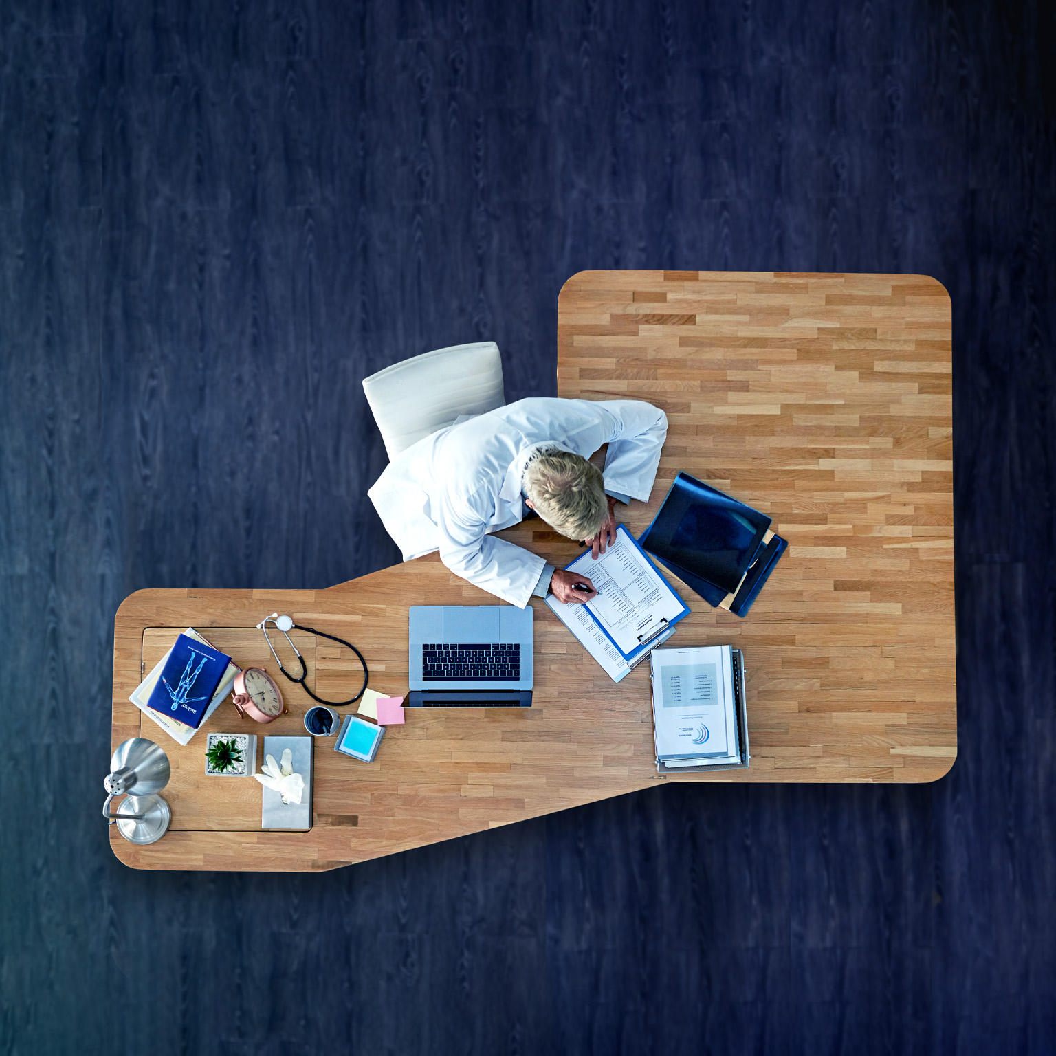 A bird's eye perspective of a physician sitting at an executive desk, filling out paperwork. Beside them sits a laptop, medical records, and a stethoscope. The desk is positioned on a rich, dark wooden floor.