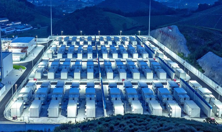 A large grid of containerized battery storage units arranged in neat rows within a brightly lit industrial facility at dusk. The site is surrounded by hills and vegetation, suggesting a large-scale energy storage installation set in a rural or semi-rural landscape.