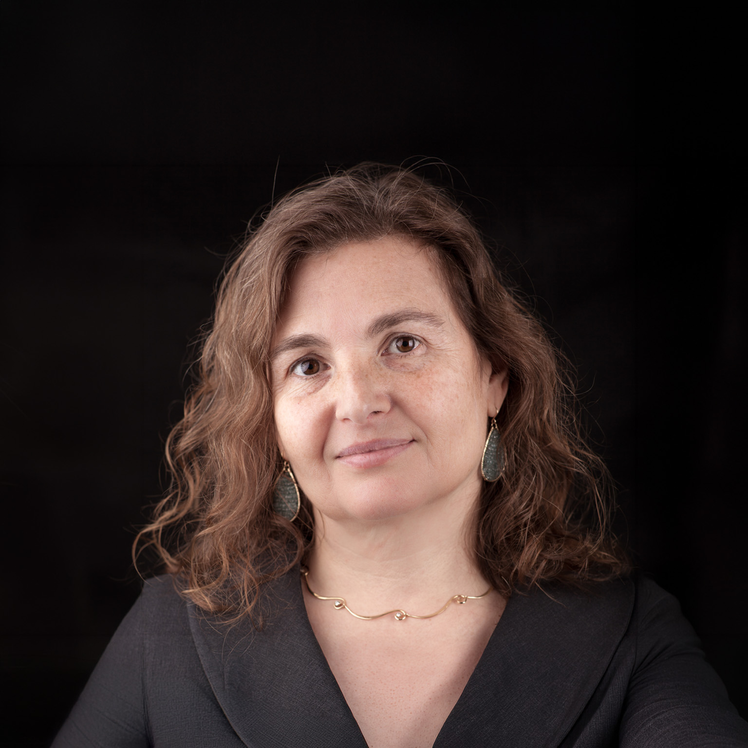 A close-up portrait of Daniela Rus with wavy brown hair, wearing a dark top and subtle jewelry against a black background.