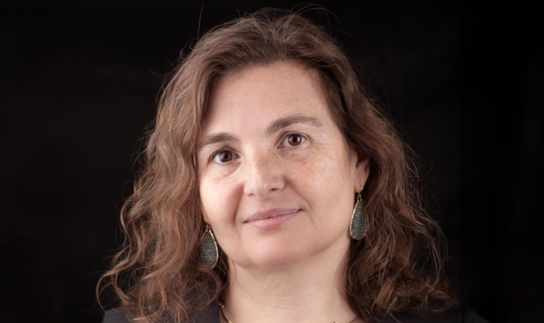A close-up portrait of Daniela Rus with wavy brown hair, wearing a dark top and subtle jewelry against a black background.