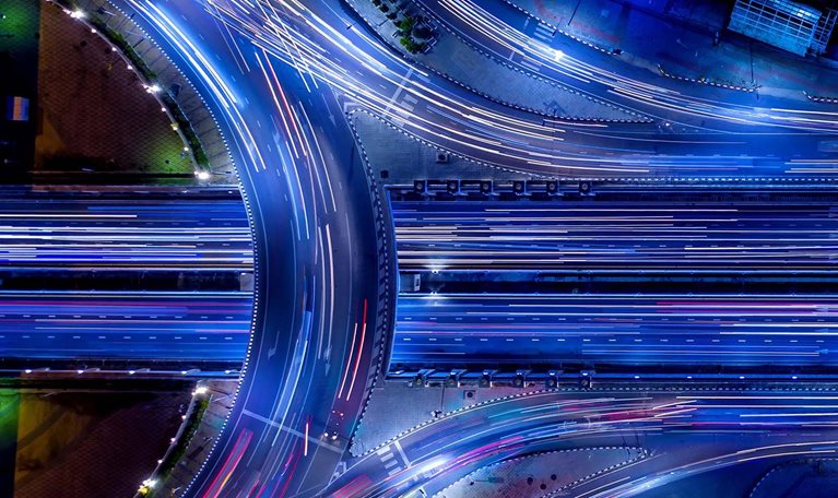 Aerial night view of a highway interchange with vibrant blue-toned long-exposure light trails from moving traffic.