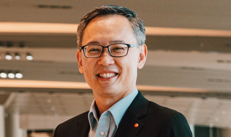 Lim Ching Kiat smiles while standing in an airport, dressed in a suit and wearing glasses.