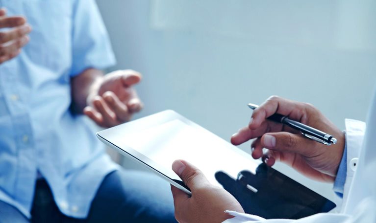 A close up of a doctor's hands holding a tablet while talking to a patient about his treatment plan.