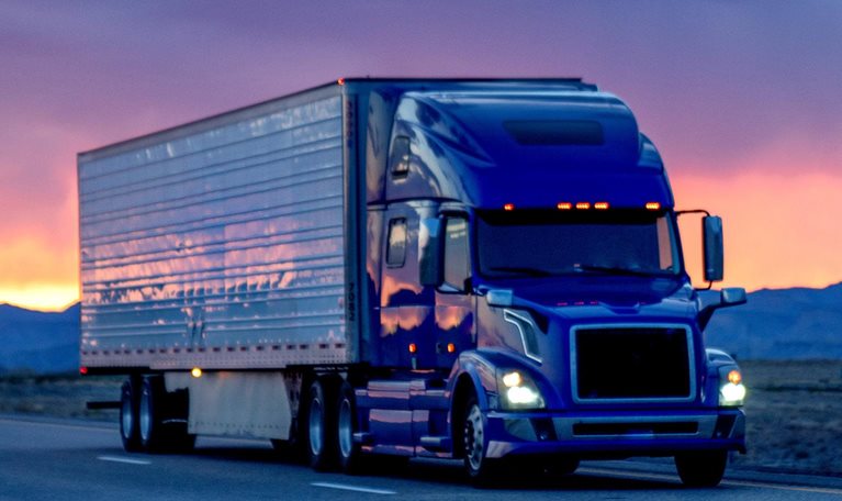 Blue semi-truck driving on a highway at sunset with cloudy sky in the background