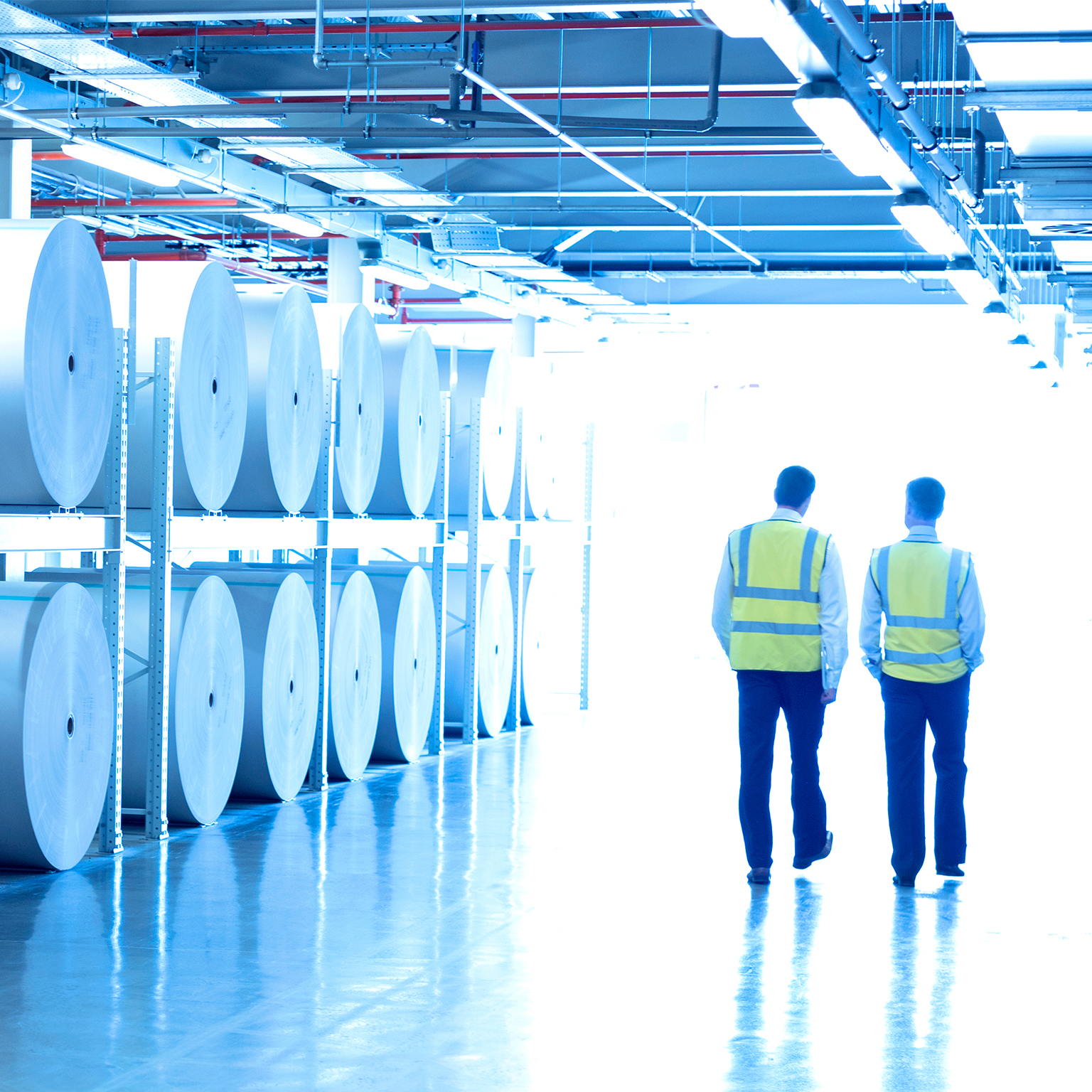 Workers in reflective clothing walking along large paper spools in printing plant