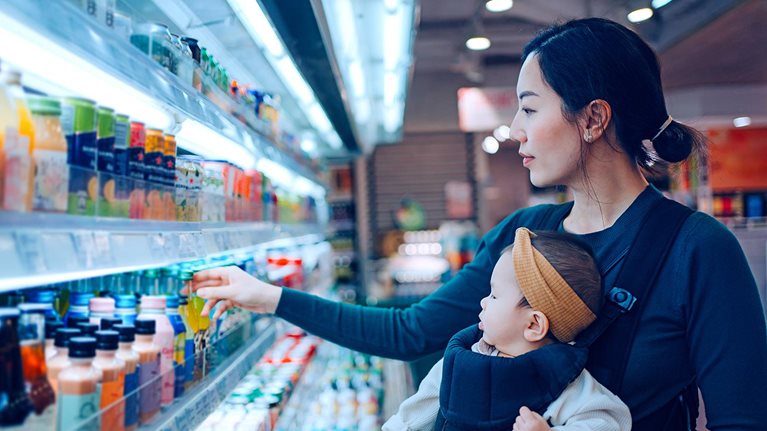 Young Asian mother carrying her baby girl in a baby carrier, doing grocery shopping in supermarket. Choosing fresh organic fruit juice for her family. Healthy eating habits with balanced diet.