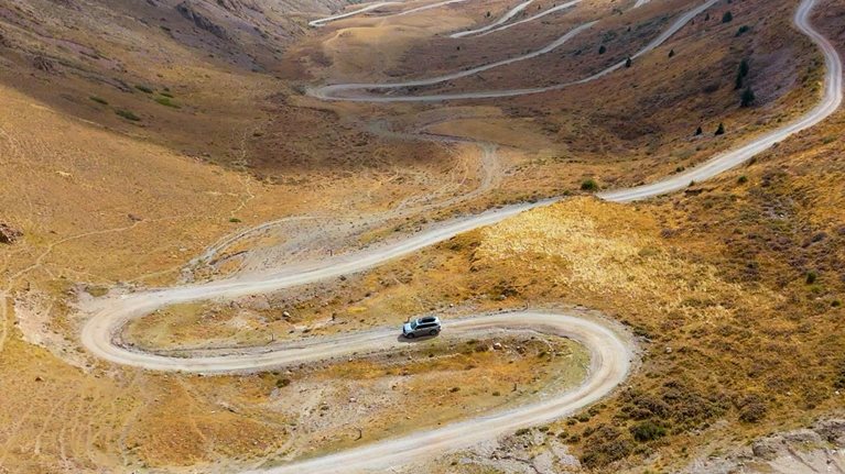 A silver SUV makes navigates a winding dirt road as it snakes through a wide mountain valley, cutting sharp switchbacks across dry, golden-brown slopes. Steep ridges rise on both sides, with sparse vegetation and distant peaks fading into a hazy, expansive backdrop.