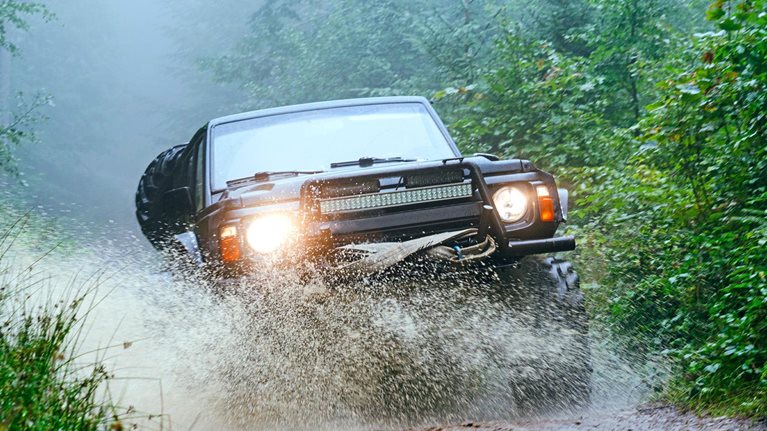 The image shows a rugged off-road vehicle driving through a muddy forest trail, splashing water as it moves forward. Dense green foliage surrounds the path, and mist hangs in the air, creating a dramatic and adventurous atmosphere.