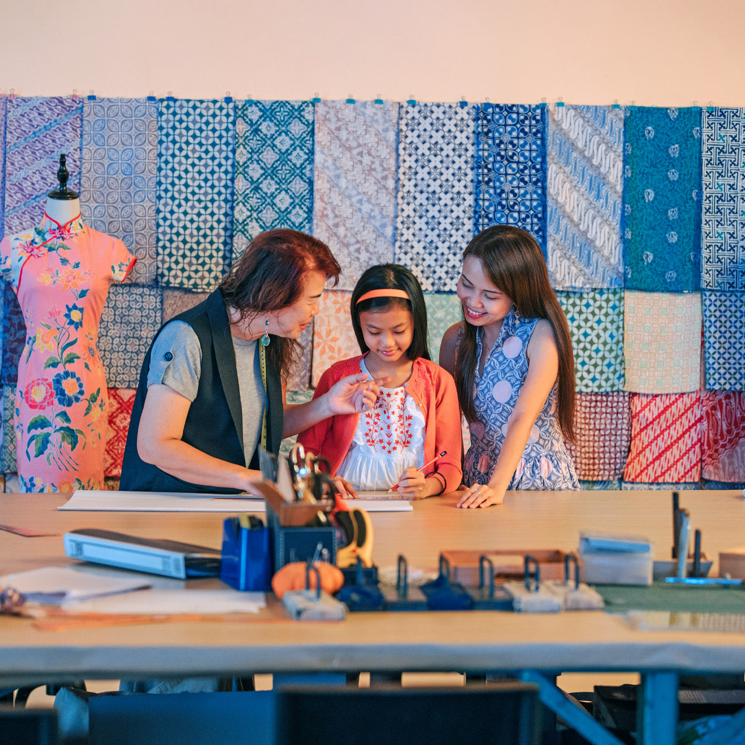 A Chinese grandmother teaching her granddaughter how to design a garment while her mother looks on. Fabric patterns hang on the wall behind them and traditional Chinese dresses hang on mannequins.  