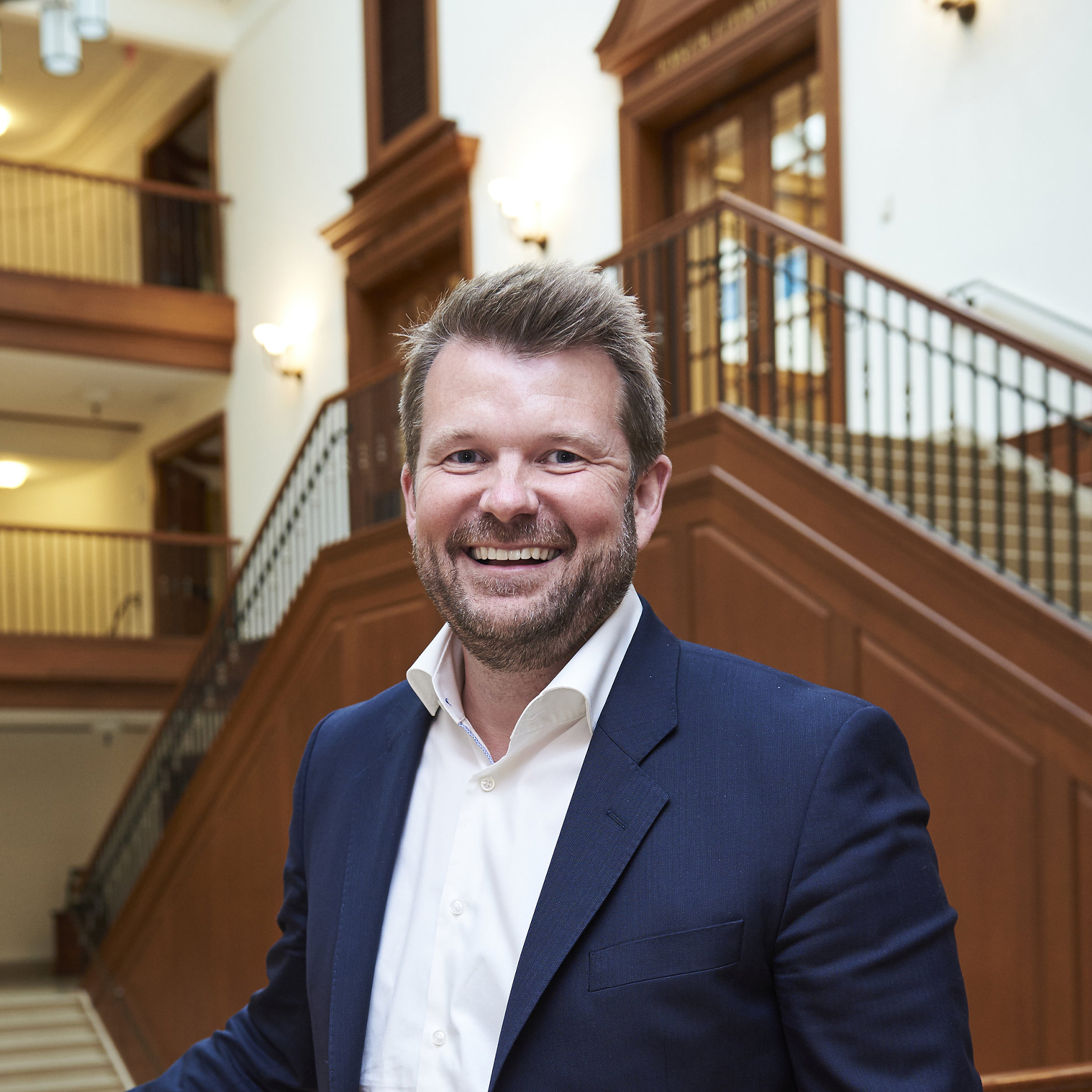 A portrait of Reynir Indahl, founder and managing partner of Summa Equity, in a dark blue suit standing in a grand, wood-paneled building with a large staircase in the background. He is leaning on a railing, looking directly at the camera.