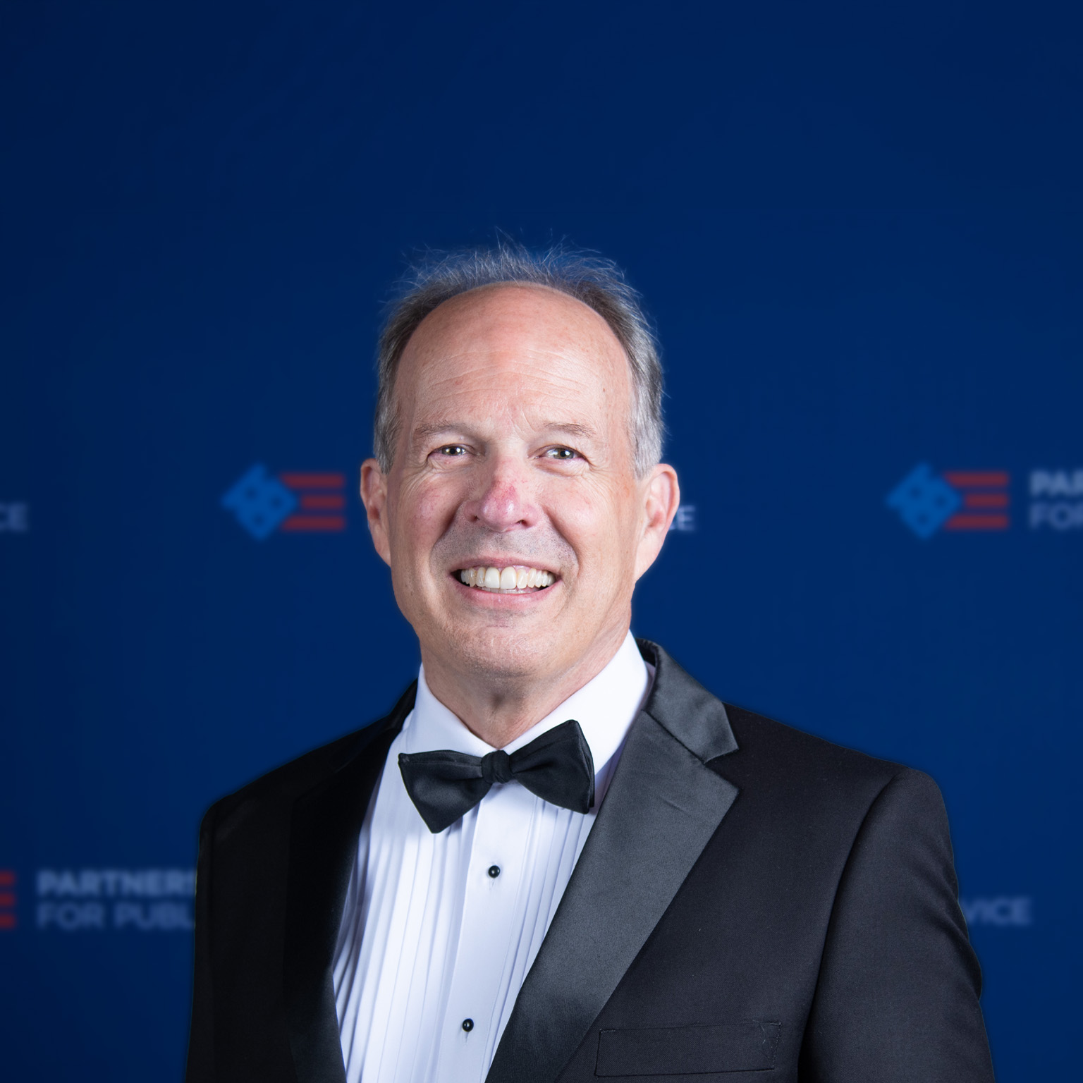 David Lebryk wearing a black tuxedo and bow tie smiles at the camera as he stands in front of a dark blue backdrop with faint logos.