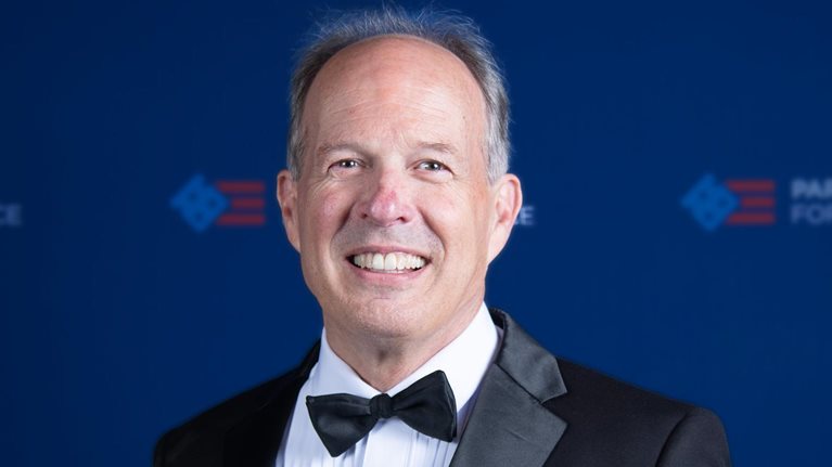 David Lebryk wearing a black tuxedo and bow tie smiles at the camera as he stands in front of a dark blue backdrop with faint logos.