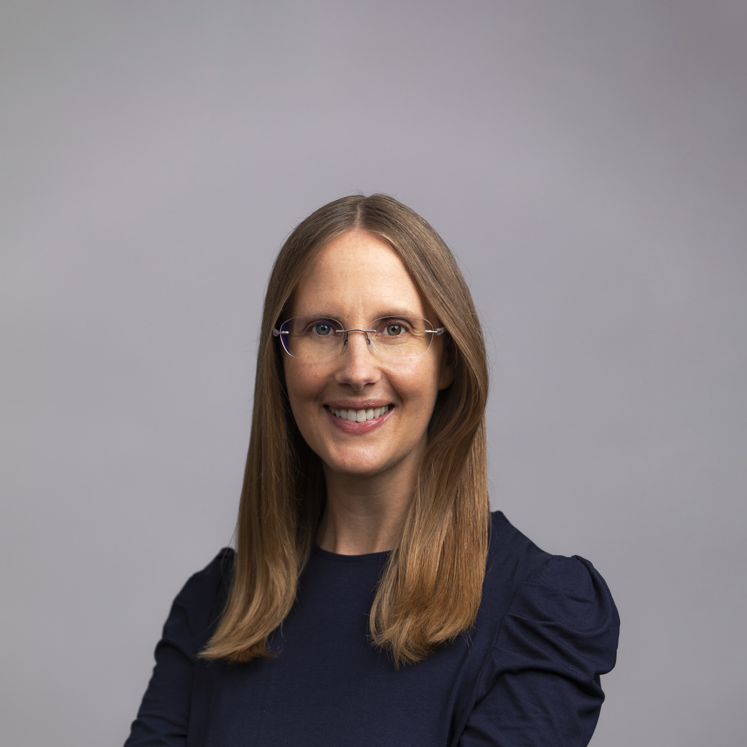 A portrait of Kelly Meissner set against a medium gray background as she sports a smile, glasses, and a navy blue blouse.