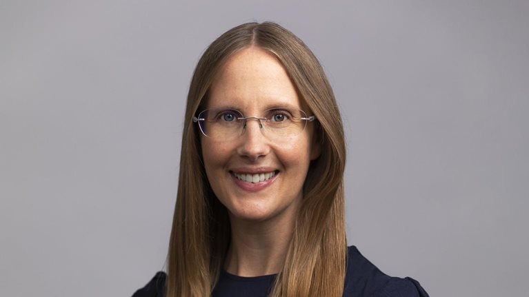 A portrait of Kelly Meissner set against a medium gray background as she sports a smile, glasses, and a navy blue blouse.